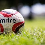 A close-up of a Mitre soccer ball resting on a grassy field with a blurred background.