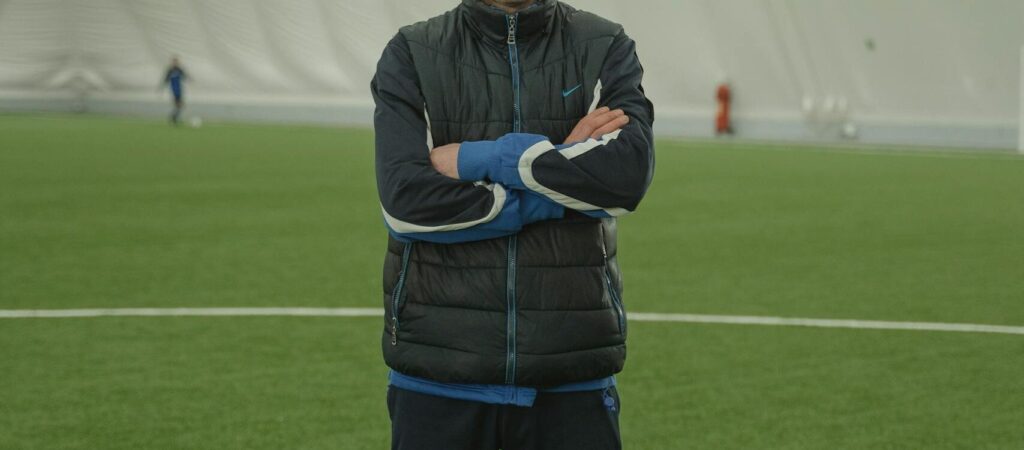 A focused soccer coach in a puffer jacket standing confidently inside an indoor stadium.
