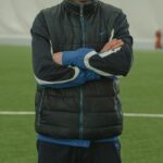 A focused soccer coach in a puffer jacket standing confidently inside an indoor stadium.