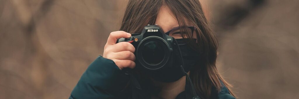 A woman with camera and face mask in a forest setting, capturing nature.